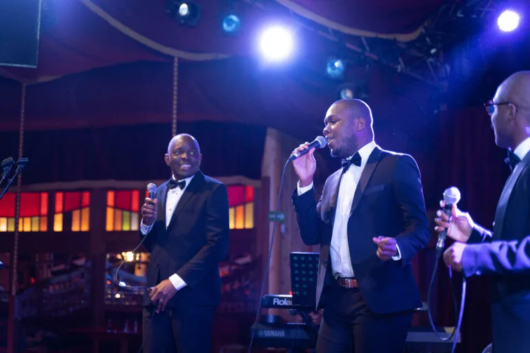 Fran's Orchestra, trois chanteurs en smoking sur scène, micro en main, interprétant une chanson sous un éclairage bleu et violet.