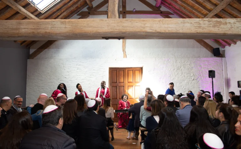 Amany Neshama, des membres d'une chorale gospel hébraïque vêtus de toges rouges et blanches chantent devant un public attentif dans une salle lumineuse avec un toit en bois.