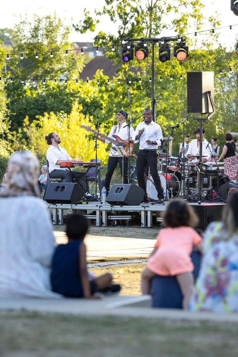 Fran's Orchestra, groupe de musiciens jouant sur une scène en plein air, avec un chanteur, un bassiste, un pianiste et un batteur, tandis que des spectateurs assis sur l’herbe profitent du concert.