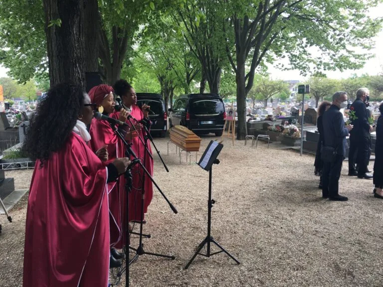 Amany, trois chanteuses gospel en robes rouges chantant lors d’une cérémonie funéraire en extérieur, devant un cercueil et des proches en deuil.