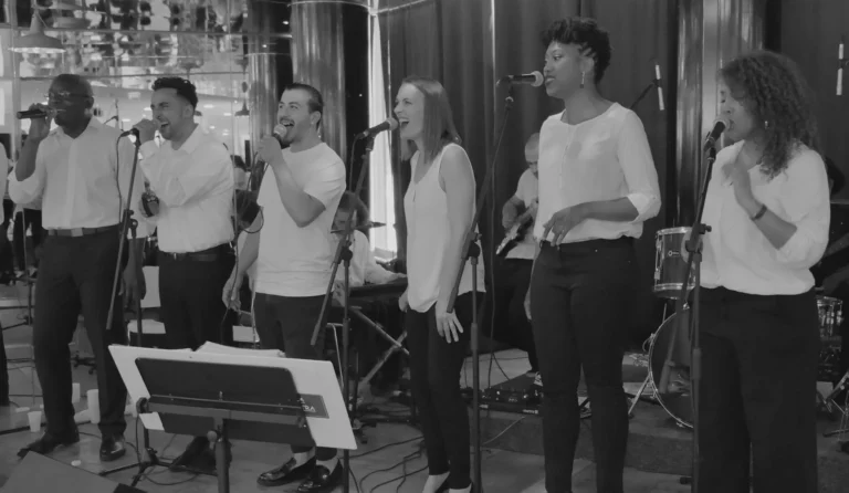 Fran's orchestra, un groupe de chanteurs gospel, vêtus de noir et blanc, interprète un morceau entraînant lors d’un concert. Leurs expressions reflètent l’émotion et l’énergie du moment, tandis que les musiciens en arrière-plan ajoutent une dimension rythmique captivante. L’ambiance est chaleureuse et immersive, propulsant le public dans une expérience musicale inoubliable.