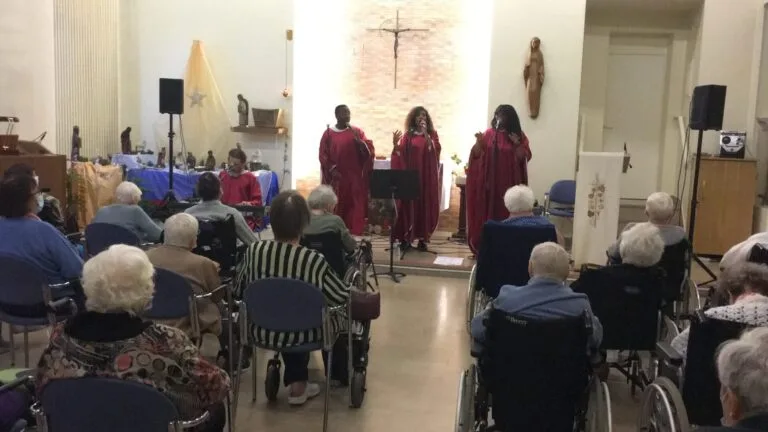 Amany, groupe de chanteurs gospel en robes rouges chantant dans une maison de retraite devant un public de résidents attentifs, certains en fauteuil roulant.