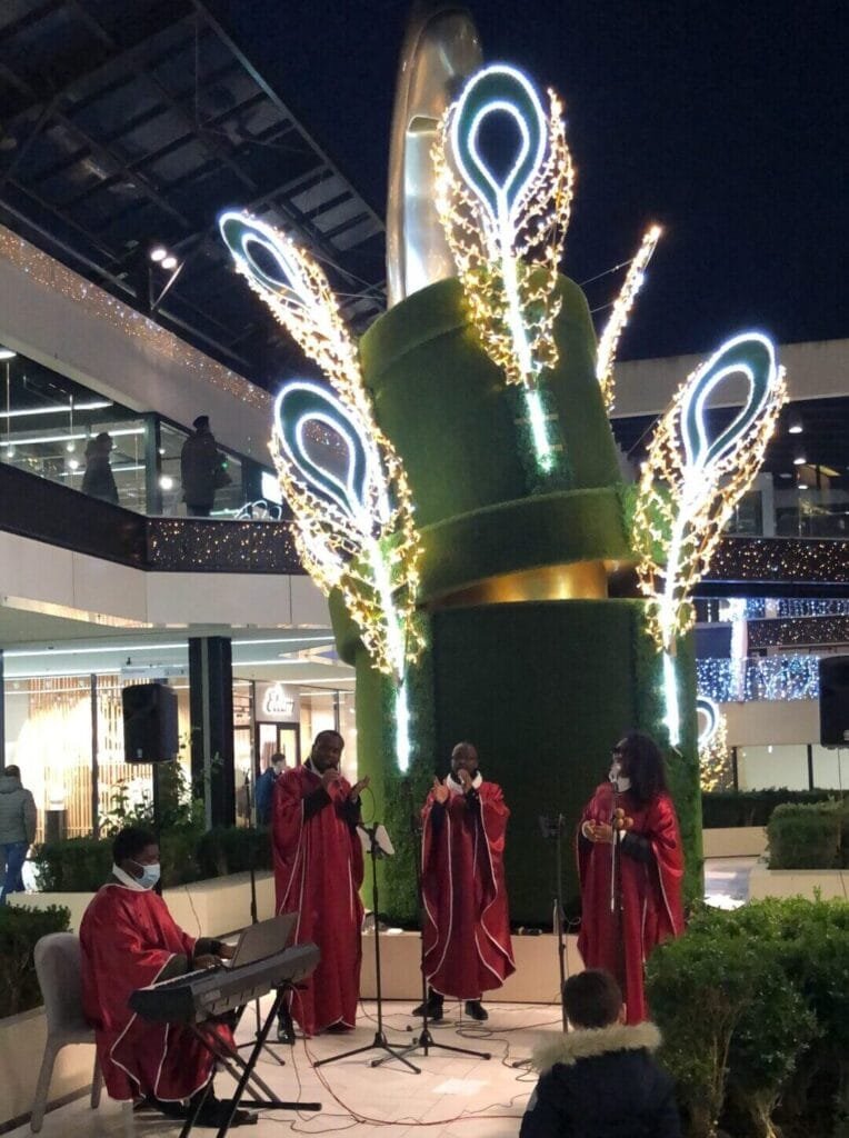 Groupe de chanteurs gospel en robes rouges interprétant des chants sous des illuminations festives dans un centre commercial, avec un pianiste et un jeune spectateur attentif.