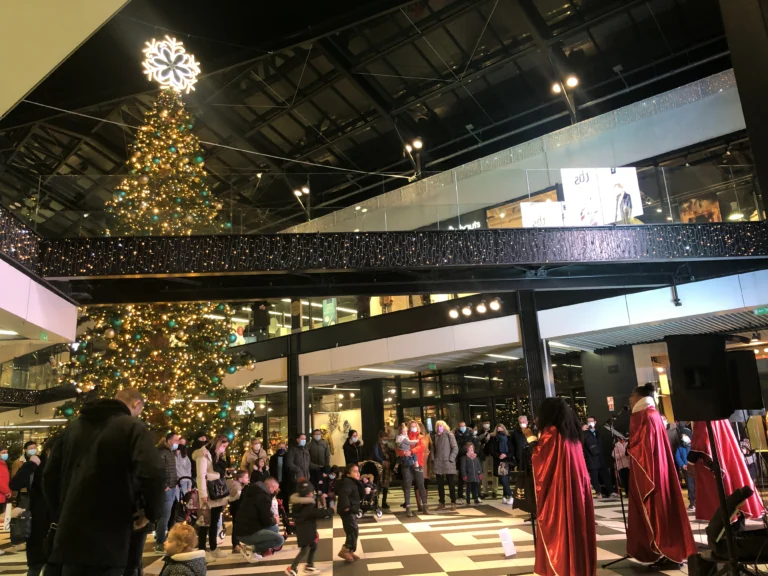 Amany, chanteurs gospel en toges rouges interprétant des chants de Noël sous un grand sapin illuminé dans un centre commercial, entourés d’un public familial.