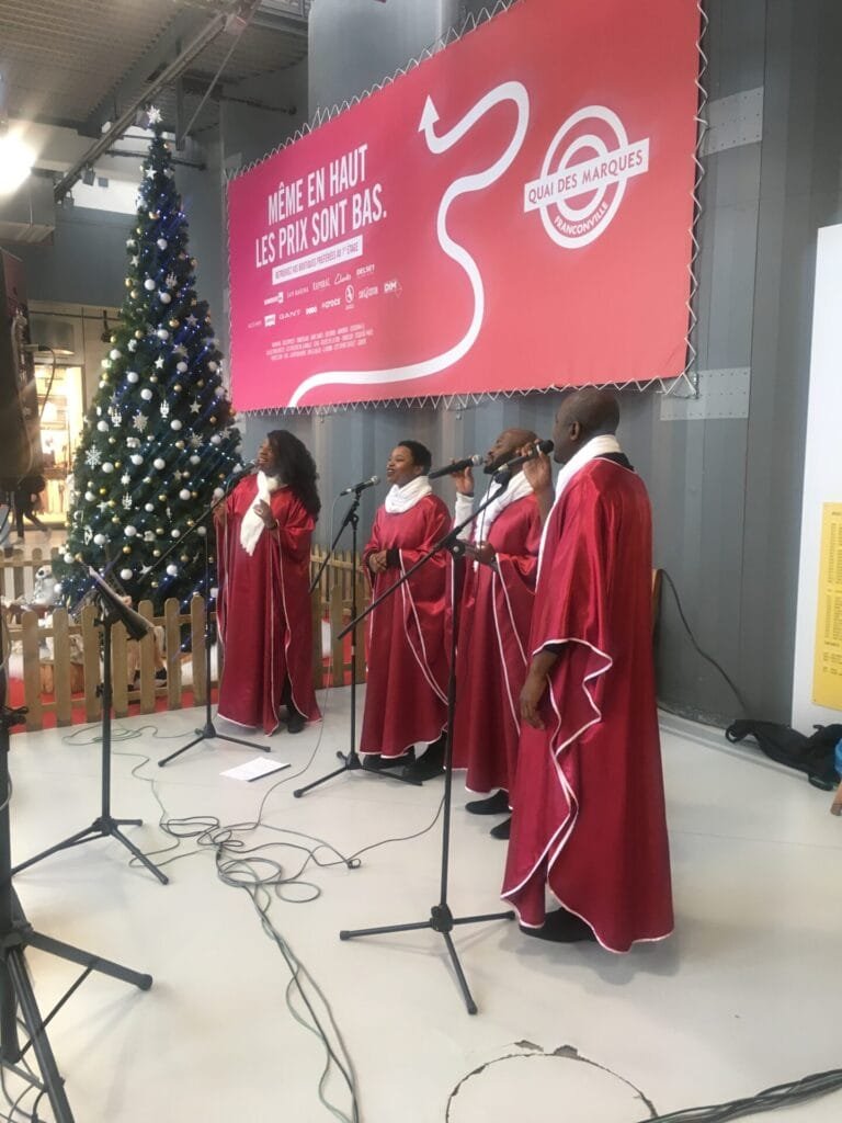 Groupe de gospel chantant dans un centre commercial décoré pour Noël, devant un grand sapin illuminé.
