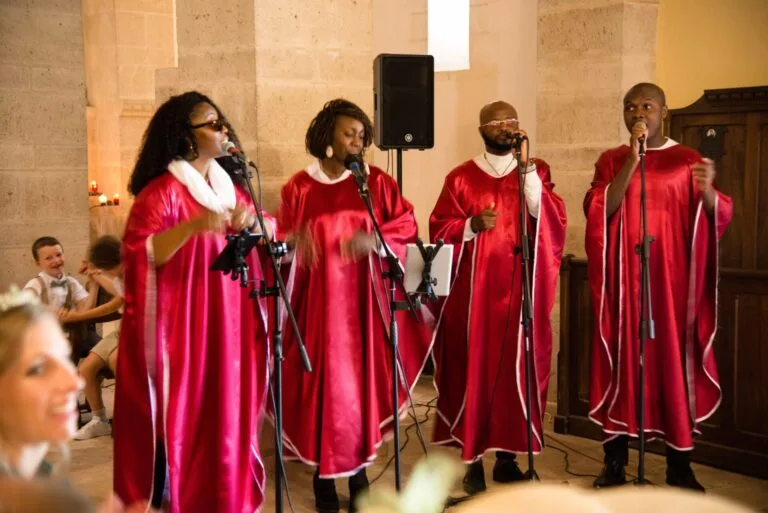 Amany, quatre chanteurs gospel en tenue rouge et blanche interprètent une chanson lors d’une cérémonie dans une église en pierre. Leur performance passionnée crée une ambiance chaleureuse et émotive, sous le regard émerveillé des invités.