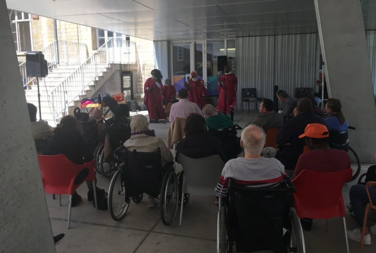Amany, concert en extérieur avec des chanteurs en robes rouges se produisant devant un public composé de résidents, dont certains en fauteuil roulant.