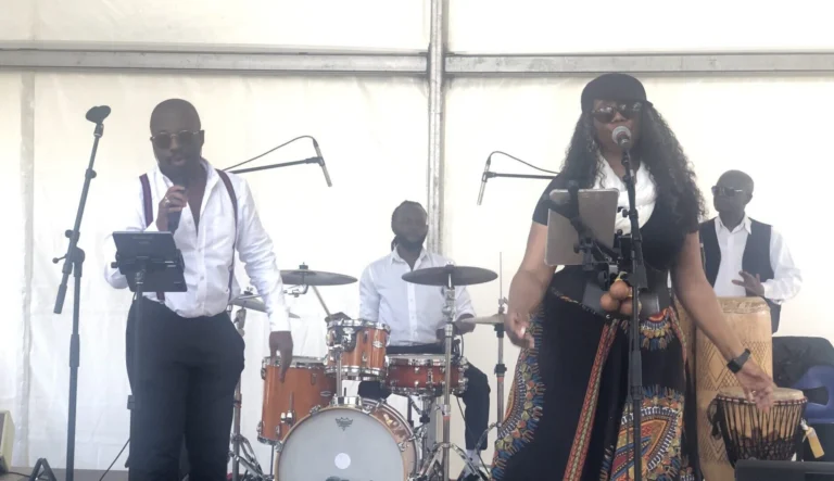 Fran's Orchestra sur scène lors d’un concert en plein air, avec une chanteuse en robe colorée, un chanteur élégant et des percussionnistes jouant en arrière-plan.