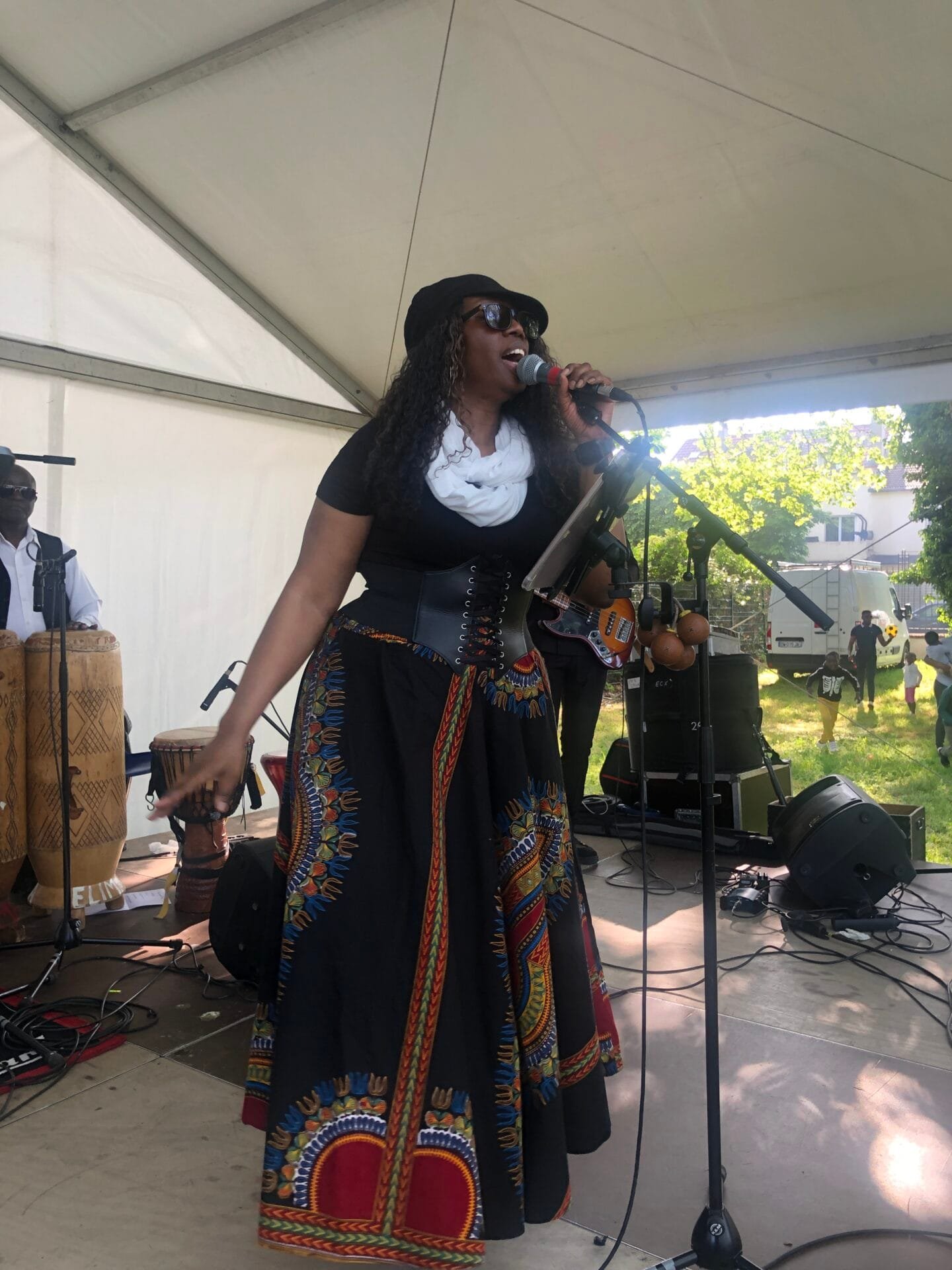 Chanteuse vêtue d'une robe colorée interprétant une chanson sur scène lors d'un festival en plein air, entourée de musiciens jouant des percussions.