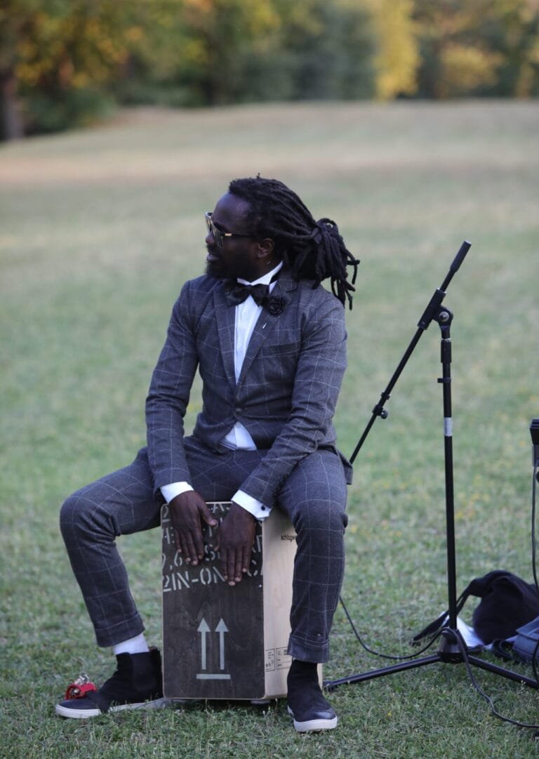 Musicien en costume jouant du cajón en extérieur, assis sur l’herbe avec un micro sur pied à proximité.