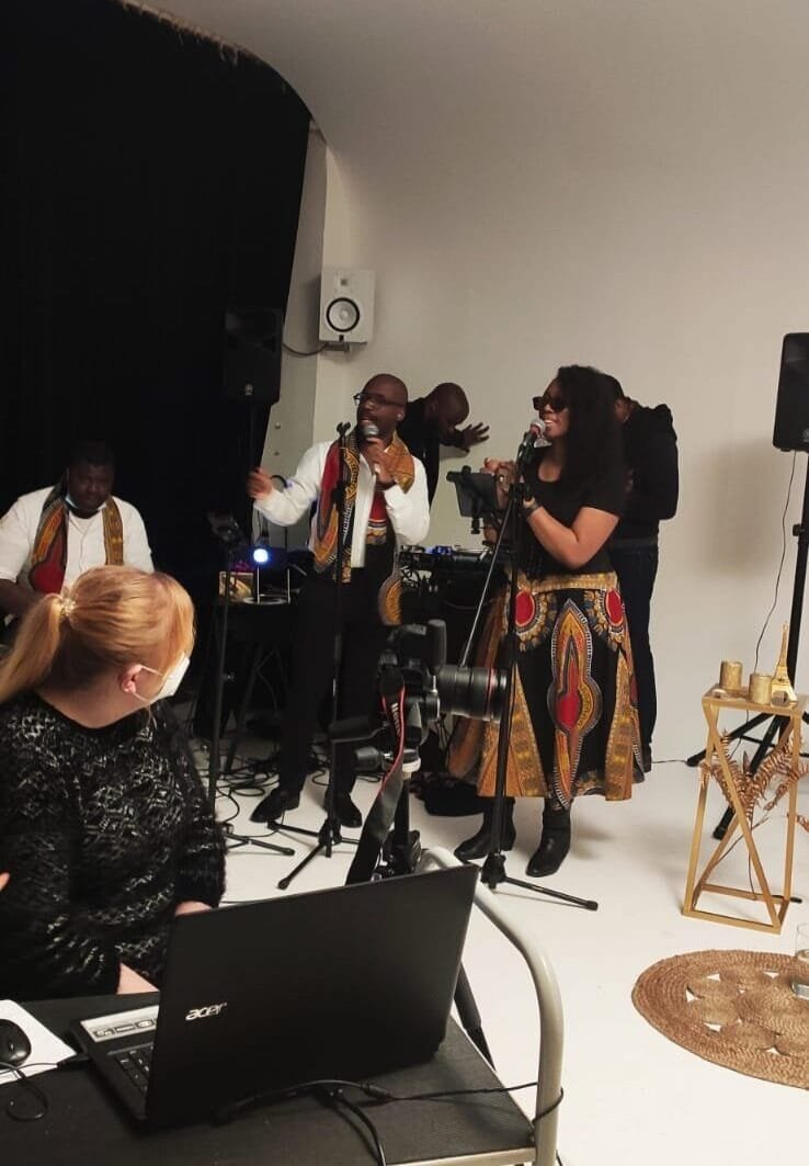 Un groupe de jazz en pleine performance live dans un studio, avec un chanteur et une chanteuse en tenues stylisées type africaines, un percussionniste et une technicienne gérant la captation de l'événement.