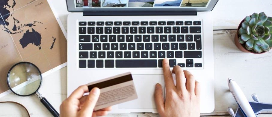Personne effectuant un achat en ligne sur un ordinateur portable, tenant une carte bancaire avec un site de voyage affiché à l'écran.
