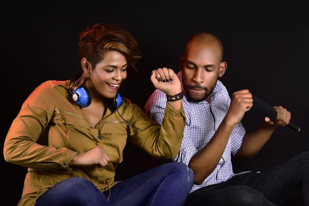 Un homme et une femme s'amusent en musique. La femme, portant un casque audio autour du cou, danse avec enthousiasme, tandis que l’homme, tenant un microphone, chante avec énergie sur un fond noir.