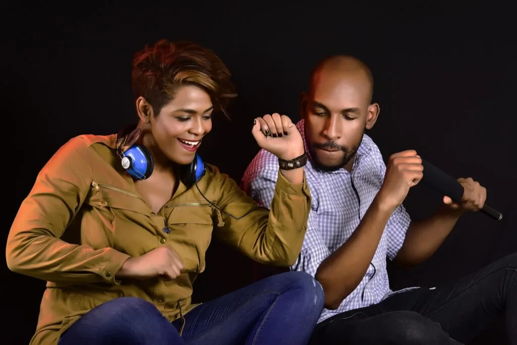 Un homme et une femme s'amusent en musique. La femme, portant un casque audio autour du cou, danse avec enthousiasme, tandis que l’homme, tenant un microphone, chante avec énergie sur un fond noir.
