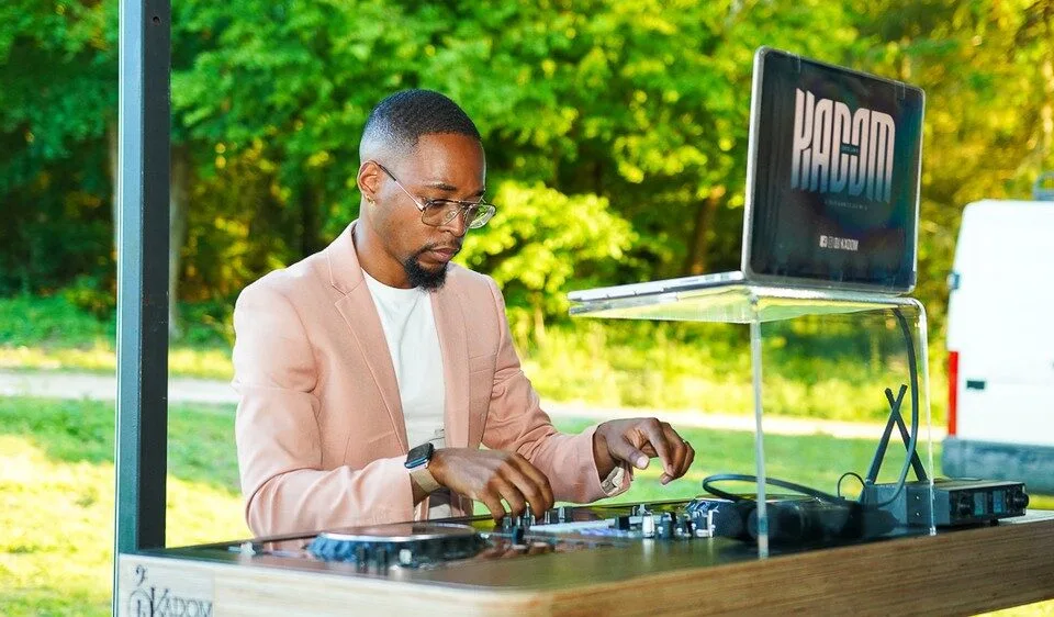 DJ Kadom, en blazer rose pâle, mixant sur ses platines en extérieur avec un ordinateur portant son nom, dans un cadre naturel et lumineux.
