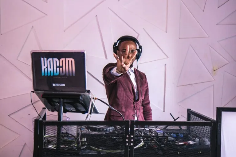 DJ Kadom en costume bordeaux, casque sur les oreilles, mixant derrière ses platines avec un ordinateur affichant son nom, dans un cadre moderne et festif.