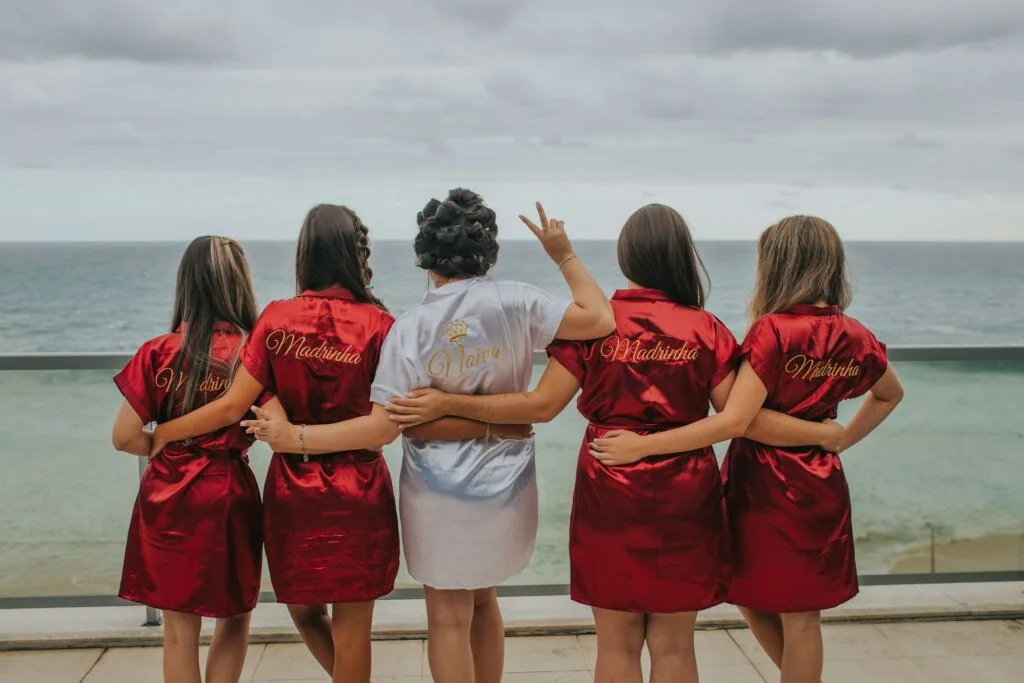 Cinq femmes sont debout sur une terrasse surplombant l'océan, habillées de robes en satin. La femme au centre, portant une robe blanche avec l'inscription 'Noiva' (mariée), est entourée de ses amies ou demoiselles d'honneur vêtues de rouge avec l'inscription 'Madrinha' (marraine ou demoiselle d'honneur). Elles s'enlacent par la taille, tournées vers la mer, tandis que la mariée fait un signe de paix avec sa main. Une scène capturant la complicité et la joie des préparatifs d’un mariage dans un cadre idyllique.