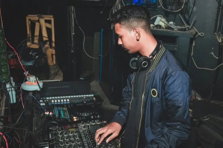 DJ concentré réglant une table de mixage dans un environnement technique avec un casque autour du cou.