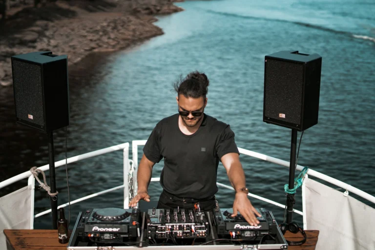 DJ en train de mixer sur un bateau, entouré de son matériel et d'une vue panoramique sur l'eau.