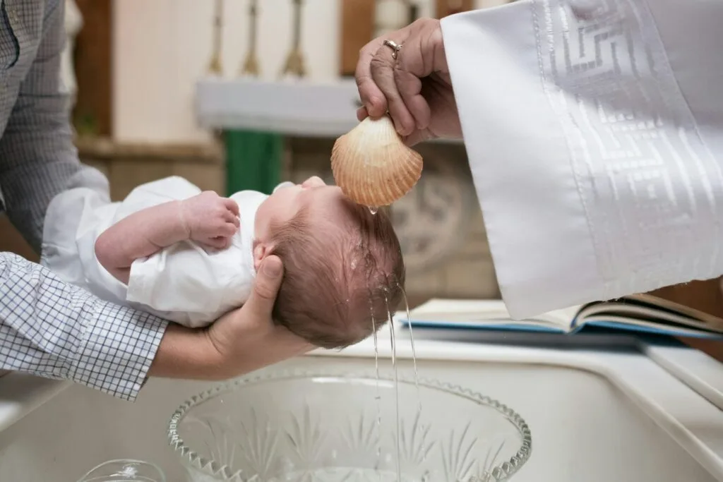 Un bébé vêtu de blanc est tenu délicatement au-dessus d'une vasque en verre tandis qu'un prêtre, vêtu d'une robe liturgique blanche, verse de l'eau sur sa tête à l'aide d'une coquille Saint-Jacques. La scène se déroule dans une église, avec un autel visible en arrière-plan, illustrant une cérémonie de baptême empreinte de spiritualité et de tradition.