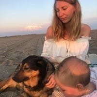 Une femme assise sur une plage au coucher du soleil, accompagnée d’un bébé et d’un chien.