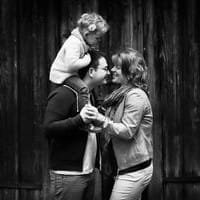 Un couple s’enlace en souriant, tandis que leur enfant, assis sur les épaules du père, touche affectueusement le front de sa mère.