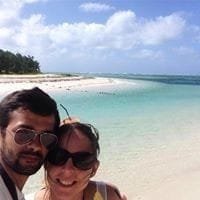 Un couple souriant prend un selfie sur une plage tropicale, avec une mer turquoise et un ciel bleu en arrière-plan.