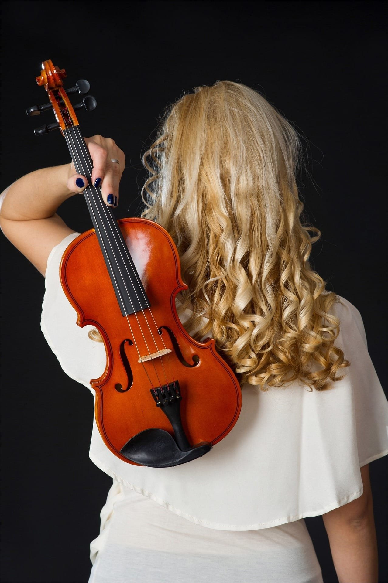 Femme aux cheveux blonds bouclés tenant un violon derrière son dos, sur fond noir.
