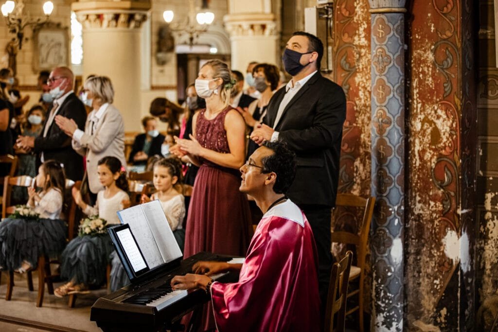 Chorale lors d'une cérémonie religieuse : un musicien en robe de chœur rose joue du clavier, entouré de participants masqués, debout et applaudissant dans une église ornée de décorations anciennes.