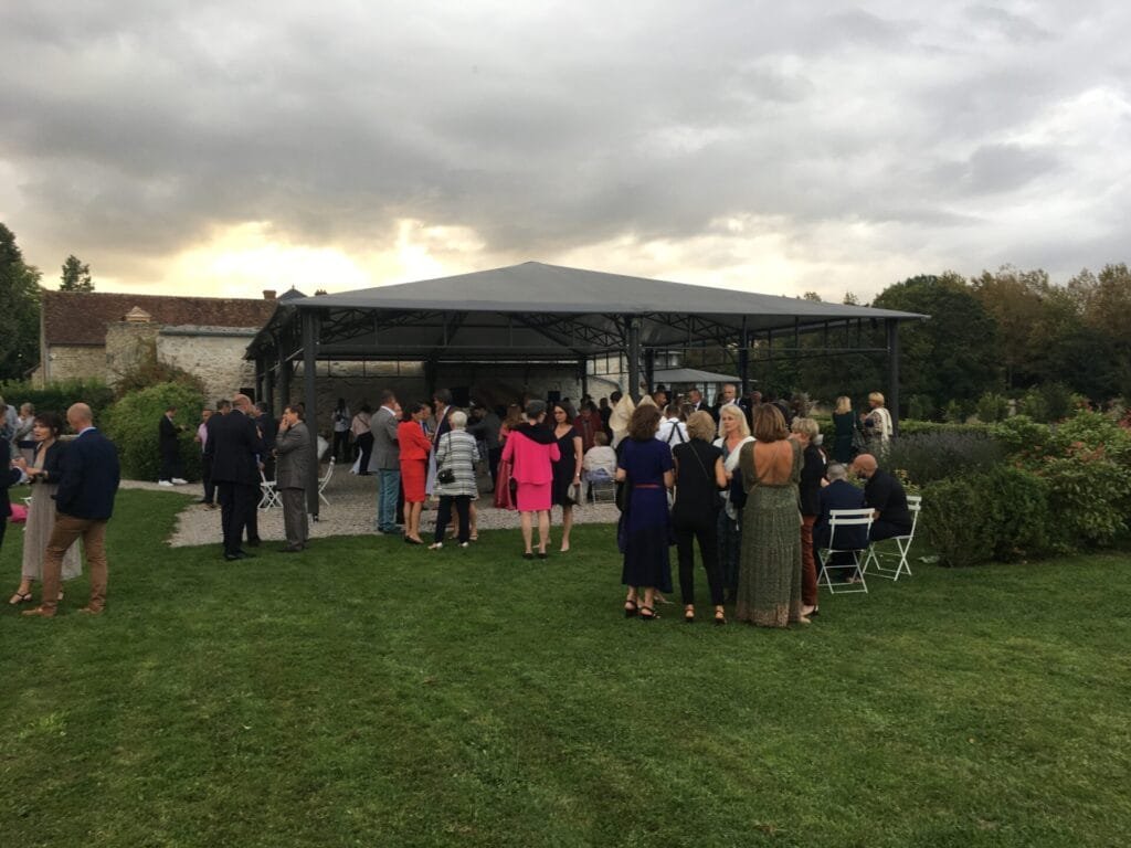 Invités réunis sous une pergola pour un cocktail de mariage élégant en extérieur, entourés de verdure et d’une bâtisse en pierre.