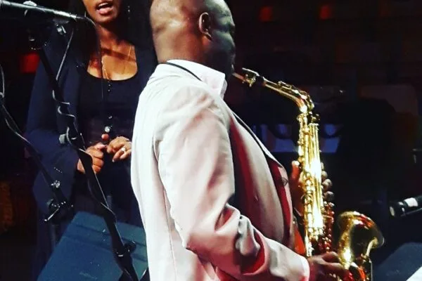 Un saxophoniste en costume blanc joue sur scène tandis qu'une chanteuse en tenue noire l'accompagne au micro.