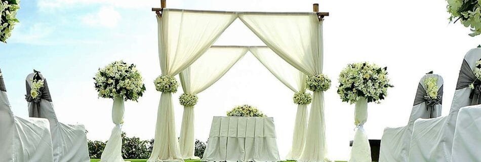 Arche de mariage en bois décorée de voilages blancs et de bouquets de fleurs blanches. Devant l’arche se trouve une table recouverte d’une nappe blanche, entourée de rangées de chaises drapées de housses blanches avec des nœuds gris et des petits bouquets floraux. Le sol est recouvert de pétales de fleurs blanches, créant une allée romantique sur l’herbe verte.