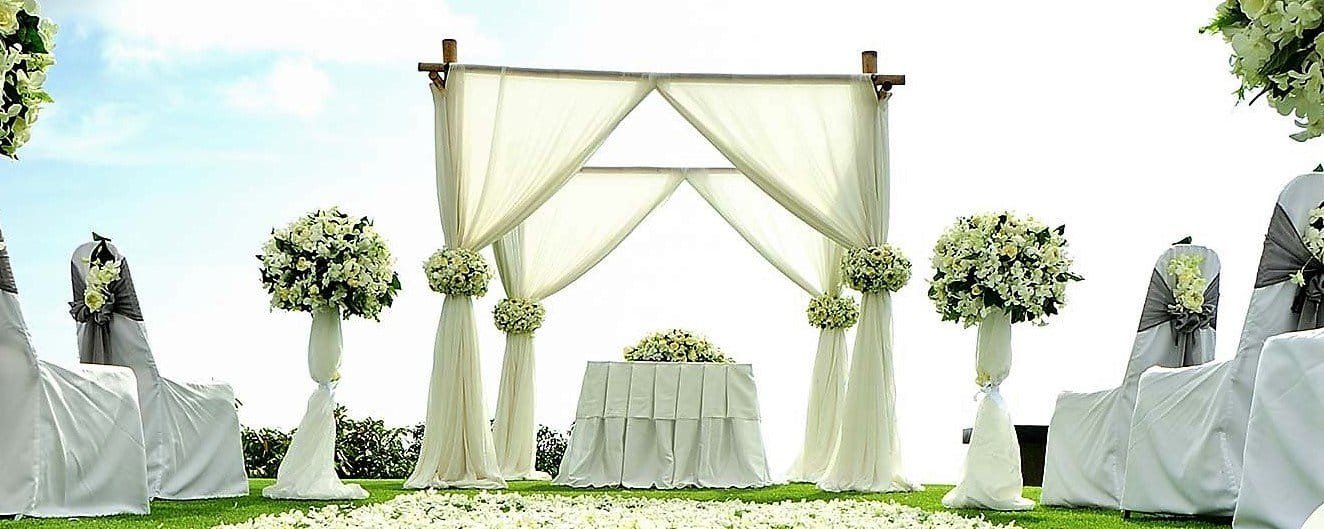 Arche de mariage en bois décorée de voilages blancs et de bouquets de fleurs blanches. Devant l’arche se trouve une table recouverte d’une nappe blanche, entourée de rangées de chaises drapées de housses blanches avec des nœuds gris et des petits bouquets floraux. Le sol est recouvert de pétales de fleurs blanches, créant une allée romantique sur l’herbe verte.