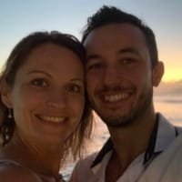 Un couple souriant prend un selfie en bord de mer au coucher du soleil.