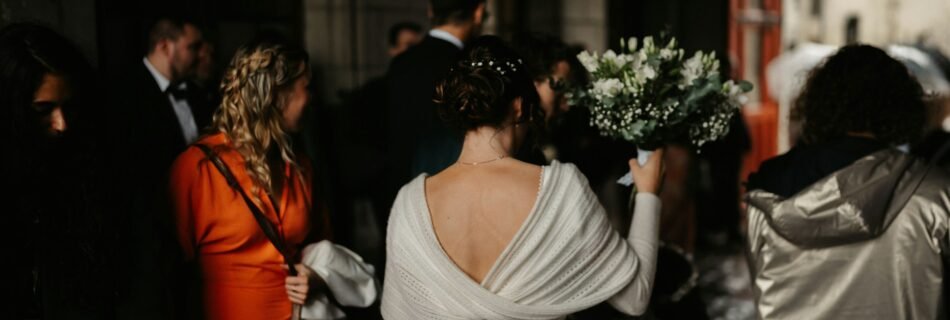 Une mariée de dos tenant son bouquet, entourée d’invités dans une ambiance chaleureuse et tamisée.