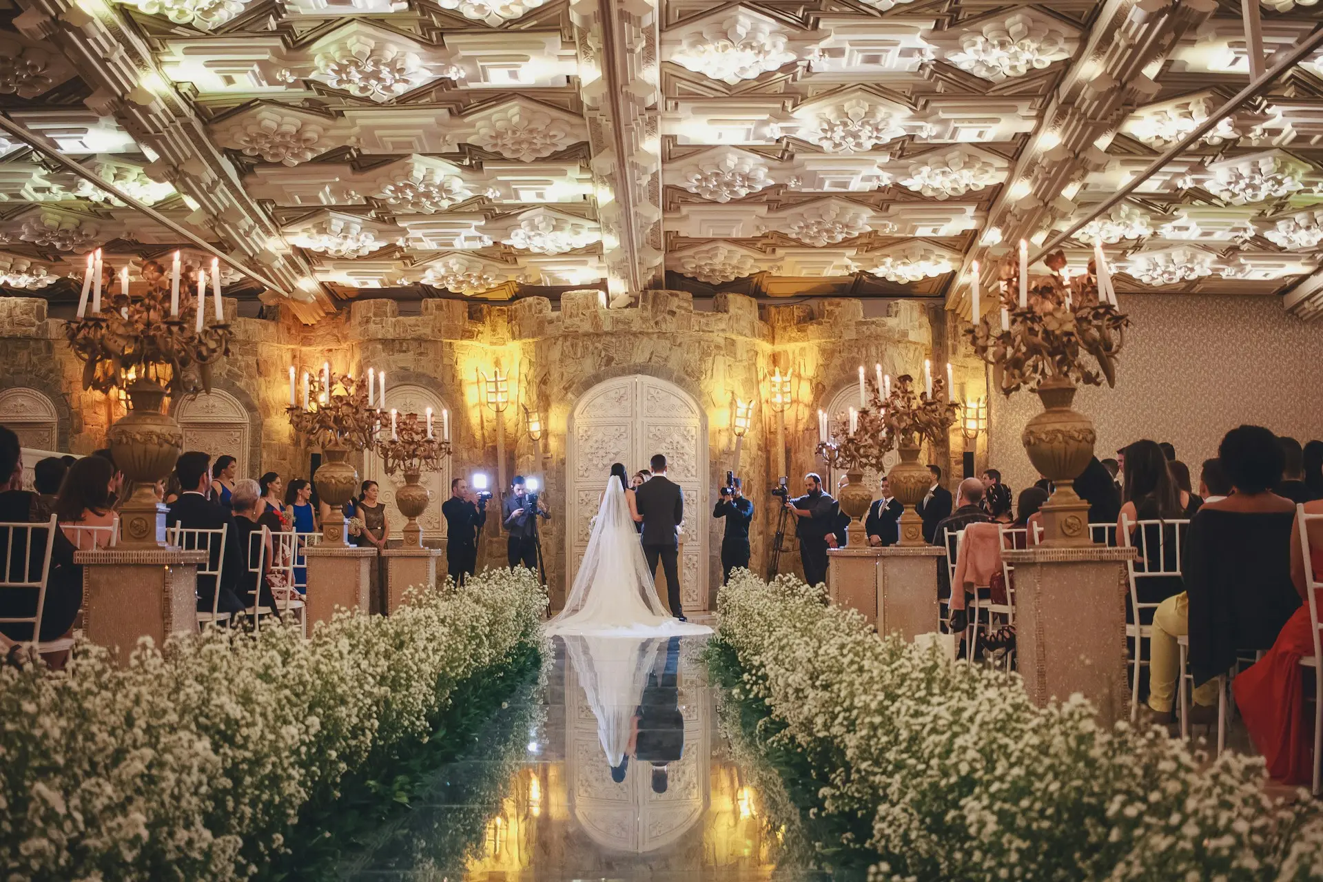 Mariage somptueux dans une salle luxueuse, avec les mariés au centre d’un décor fleuri et élégant entourés d’invités.