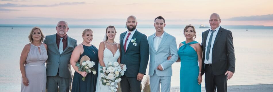 Photo de groupe d’un mariage sur la plage avec les mariés entourés de leurs proches au coucher du soleil.