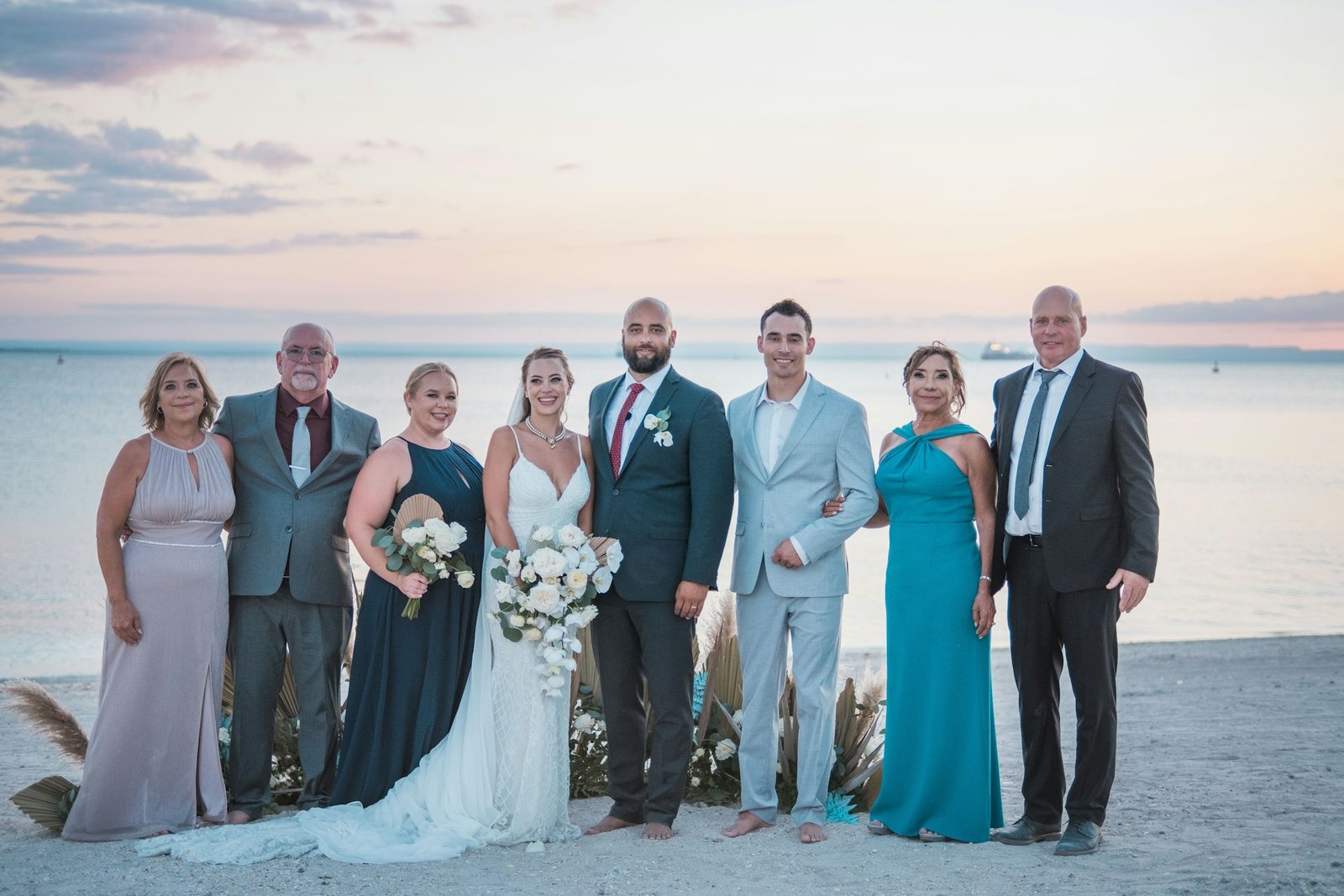 Photo de groupe d’un mariage sur la plage avec les mariés entourés de leurs proches au coucher du soleil.
