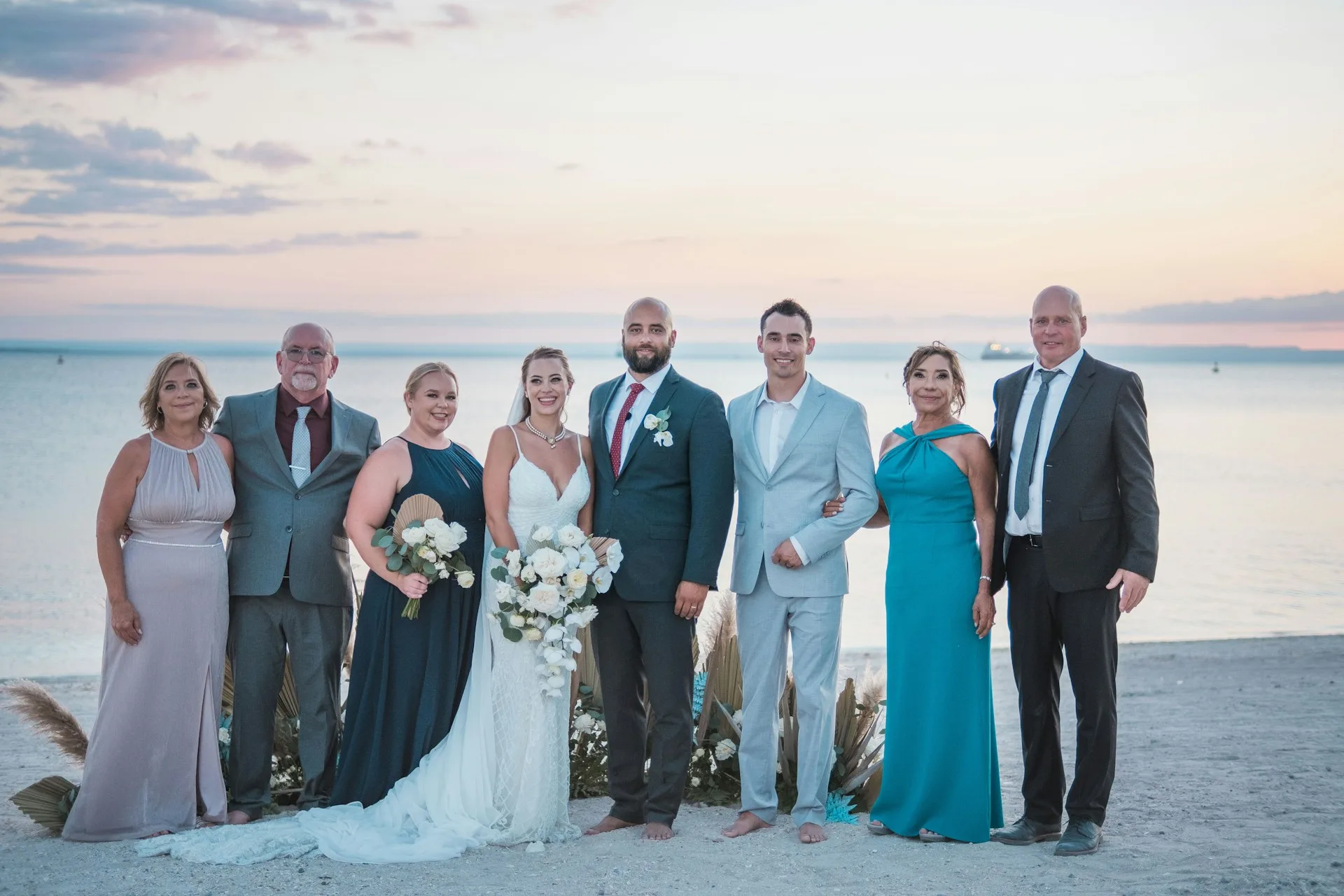 Photo de groupe d’un mariage sur la plage avec les mariés entourés de leurs proches au coucher du soleil.