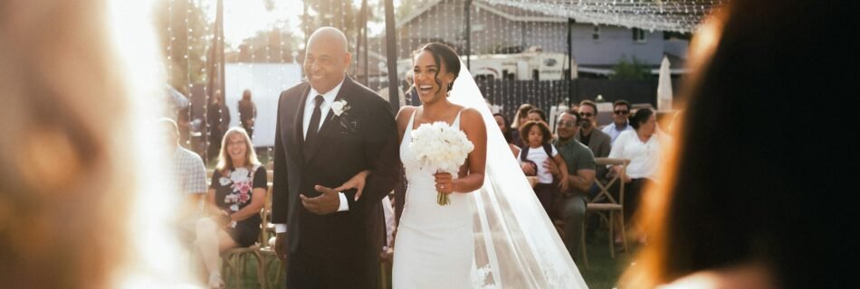 Une mariée radieuse au bras de son père avançant dans l’allée, sous le regard joyeux des invités lors d’un mariage en plein air.